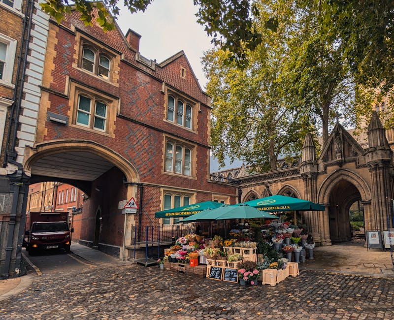 A vibrant flower stall situated on a cobbled street beneath a large green umbrella branded with flowerslee.co.uk, featuring an array of fresh flowers in various colours including pinks, reds, yellows,
