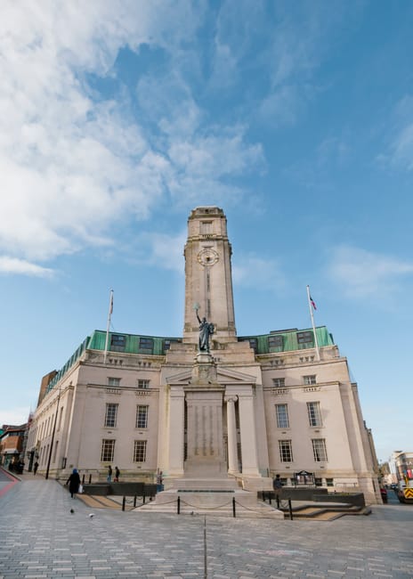 The image depicts a tall, historic monument with a central clock tower topped by a statue, set against a bright blue sky with scattered white clouds. The monument is constructed from light-colored sto