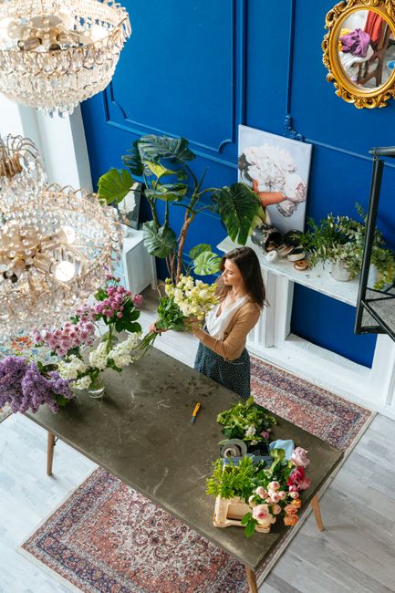 Inside a bright, elegantly decorated florist shop, a woman with long brown hair, wearing a beige top and black skirt, is arranging a fresh bouquet of flowers at a large grey worktable. The bouquet inc