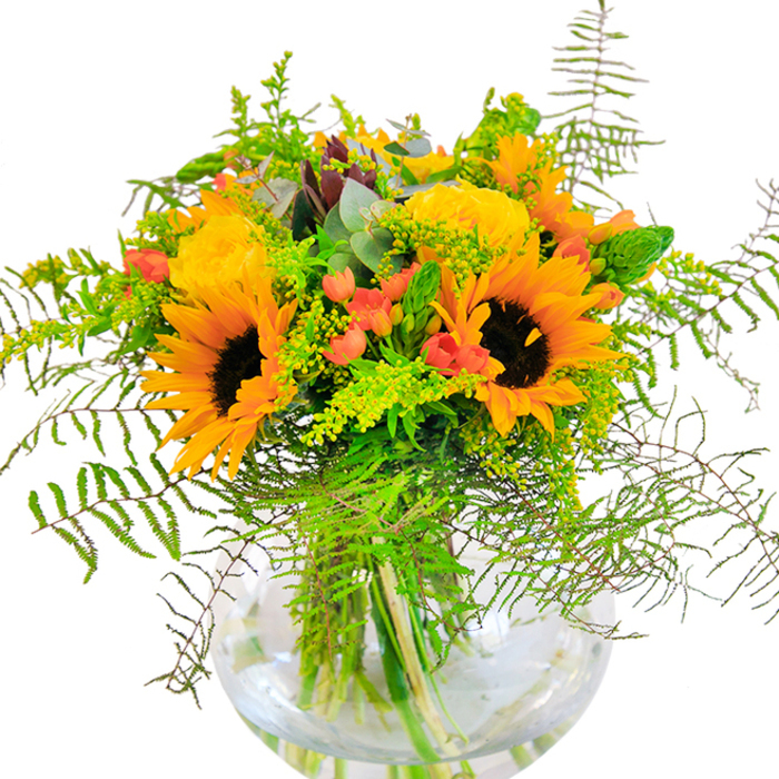 Upon first look, this stunning bouquet emits an exuberant glow-with sunlight hues of yellow and orange arranged to capture the lively character of Lee. Vibrant sunflowers, each one vivid and broad-faced, stand proudly among garden roses whose tightly coiled petals unfold in buttery layers. The crisp green of assorted foliage frames the scene, with ferns and airy sprigs gently trailing outward and lending the bouquet a dynamic sense of movement, much like the greenery found in Edith Nesbit Gardens. Pops of glossy red hypericum berries punctuate the softer shades, their sheen offering a cheerful contrast against the matte finish of the blooms, reminiscent of the vibrant market stalls found along Lee High Road. Cool eucalyptus leaves peek between flowers and light green filler plants, deepening the natural, almost wild effect of the composition. The clear, round vase not only displays the intertwining stems in fresh water but also enhances the bouquet's sense of architecture and light. In a quiet kitchen or on a busy dining table, the bouquet invites a mood of warmth and celebration-its scent subtle yet uplifting, like an English summer captured indoors. Ideal for birthdays, anniversaries, or simply to bring a touch of Lee's blooming charm into your home, this arrangement radiates happiness wherever it's placed.