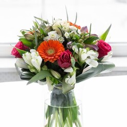 This captivating bouquet, prepared with artistic flair in a Lee flower studio, is an elegant celebration of colour and life. Prominent orange and creamy-peach gerbera daisies sit confidently at the center, their open faces shining out among a supporting cast of deep magenta roses, whose velvet whorls bring a sense of depth and romance to the arrangement. Sprays of pristine white alstroemeria mingle with airy white filler blooms, providing gentle highlights and intricate texture throughout the design. Scattered pink blossoms heighten the visual interest, while layers of lustrous greenery-especially the bold, paddle-shaped aspidistra leaves, elegantly arched around the base-frame the bouquet in a lush, comforting embrace. All are presented in a slim, transparent glass vase, where the stems' vivid greens are showcased like broad strokes on an artist's canvas. The bouquet is lit by soft, diffused daylight-perhaps reminiscent of the gentle sunshine filling a Lee cafè on Burnt Ash Road-bringing vitality to each detail. This floral arrangement perfectly captures the joyous spirit and sense of renewal unique to SE12, making it a fitting centrepiece for celebrations, intimate gatherings, or thoughtful apologies alike. With every stem positioned just so, and every colour harmoniously balanced, it's clear this design comes from hands well-versed in the tradition of local English floristry and a heartfelt dedication to the Lee community.